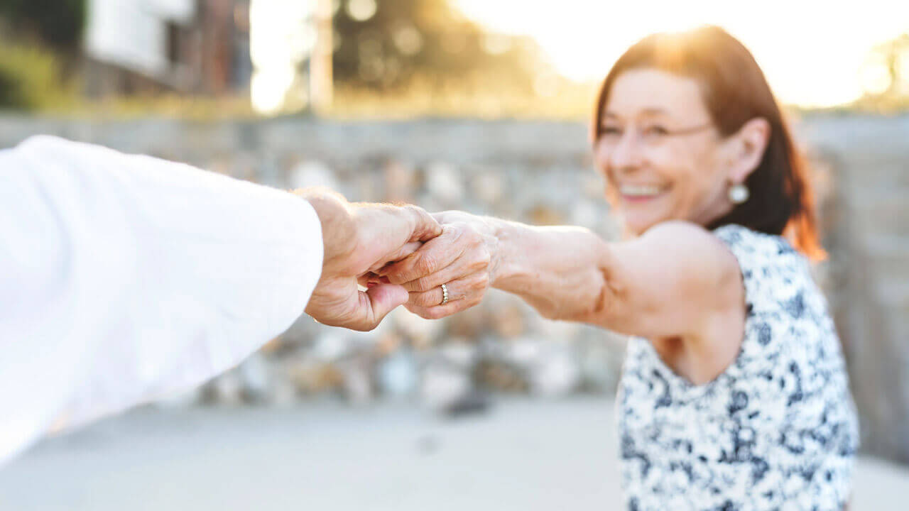 A smiling woman with dark hair and a patterned dress holds hands with another person,