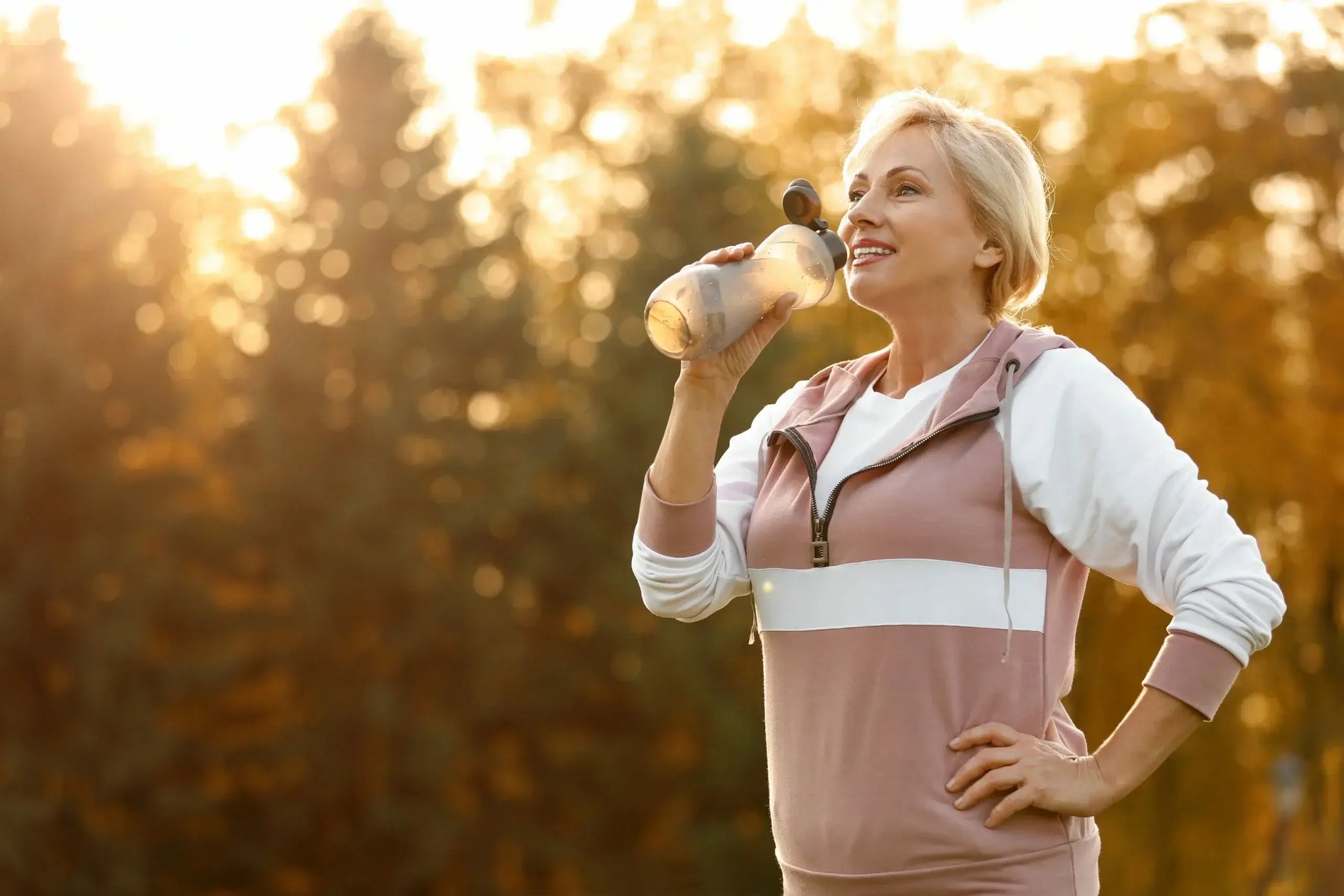A woman with short blonde hair wearing a pink and white sporty hoodie smiles while holding a clear bottle and drinking from it outdoors.