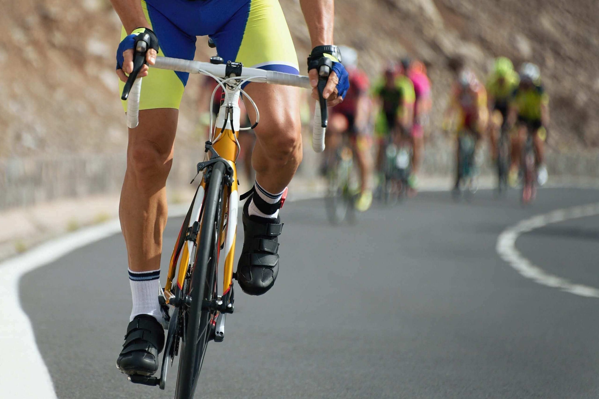 5 Great Close-up of a cyclist wearing yellow and blue spandex, black shoes, and gloves, gripping the handlebars while riding on a paved road.