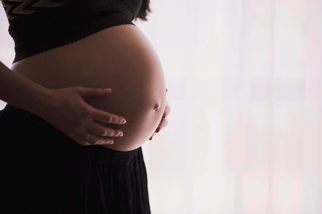 A pregnant woman gently cradles her belly with both hands. She is wearing a black skirt and a dark top.