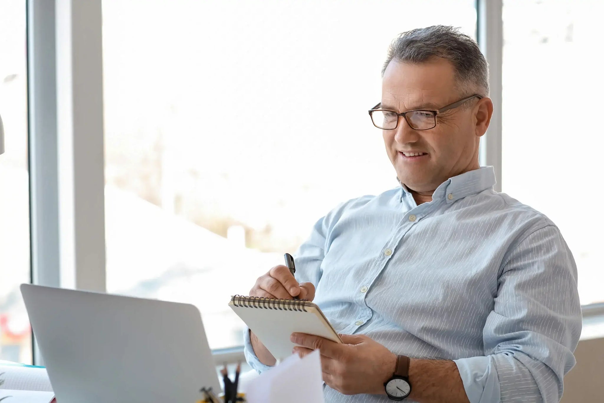 A man with short, graying hair and wearing glasses is sitting at a desk in front of a laptop.