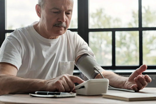 A man wearing a white t-shirt sits at a table by a large window, using a digital blood pressure monitor on his left arm.