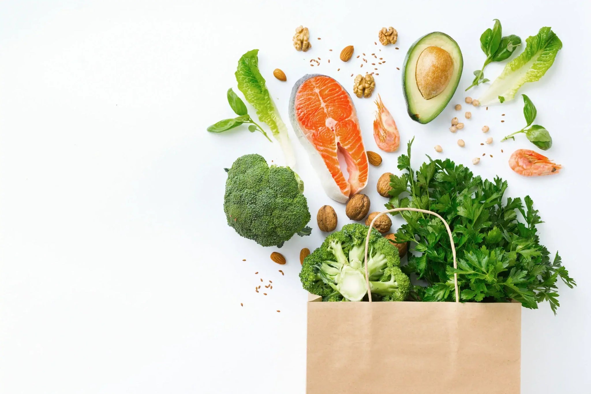 A brown paper bag on a white background, overflowing with healthy foods, including broccoli, salmon, avocado, shrimp
