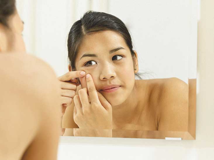 A person with dark hair is in a bathroom, looking into a mirror while touching their face, seemingly examining or popping a pimple.