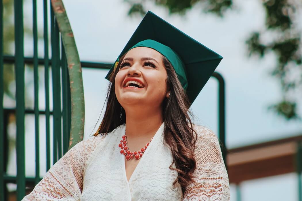 A woman in a green graduation cap and white lace dress smiles brightly while looking up, celebrating the added benefits series of her achievements.