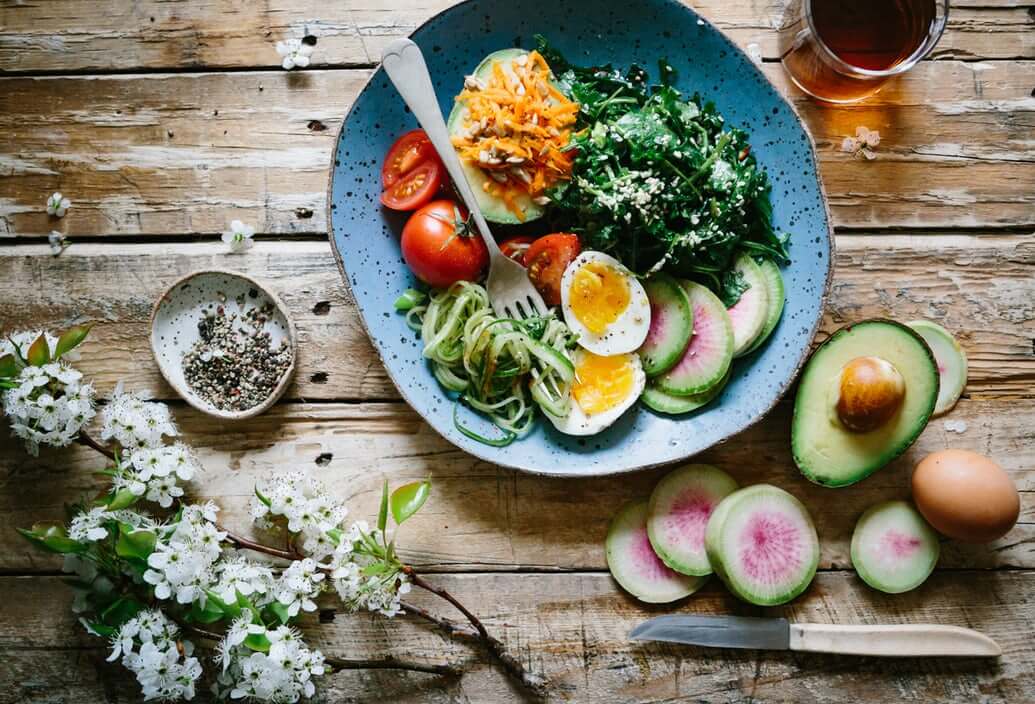 A colorful, healthy meal is presented on a rustic wooden table. The blue plate contains halved boiled eggs, sliced cucumbers, avocado, tomatoes, spiralized zucchini, grated carrots, and mixed greens.