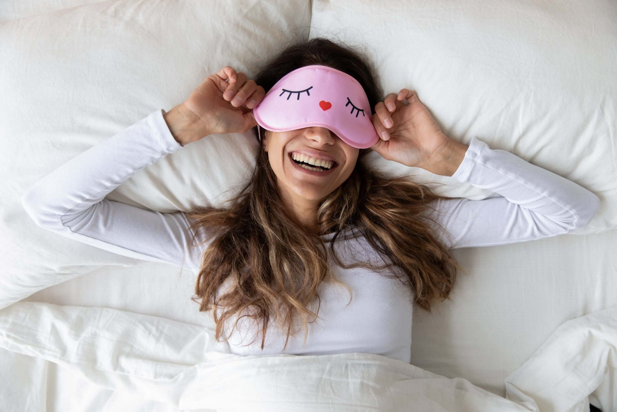 A woman is lying in bed, smiling widely while lifting a pink sleep mask decorated with cartoon eyes and a tiny heart.