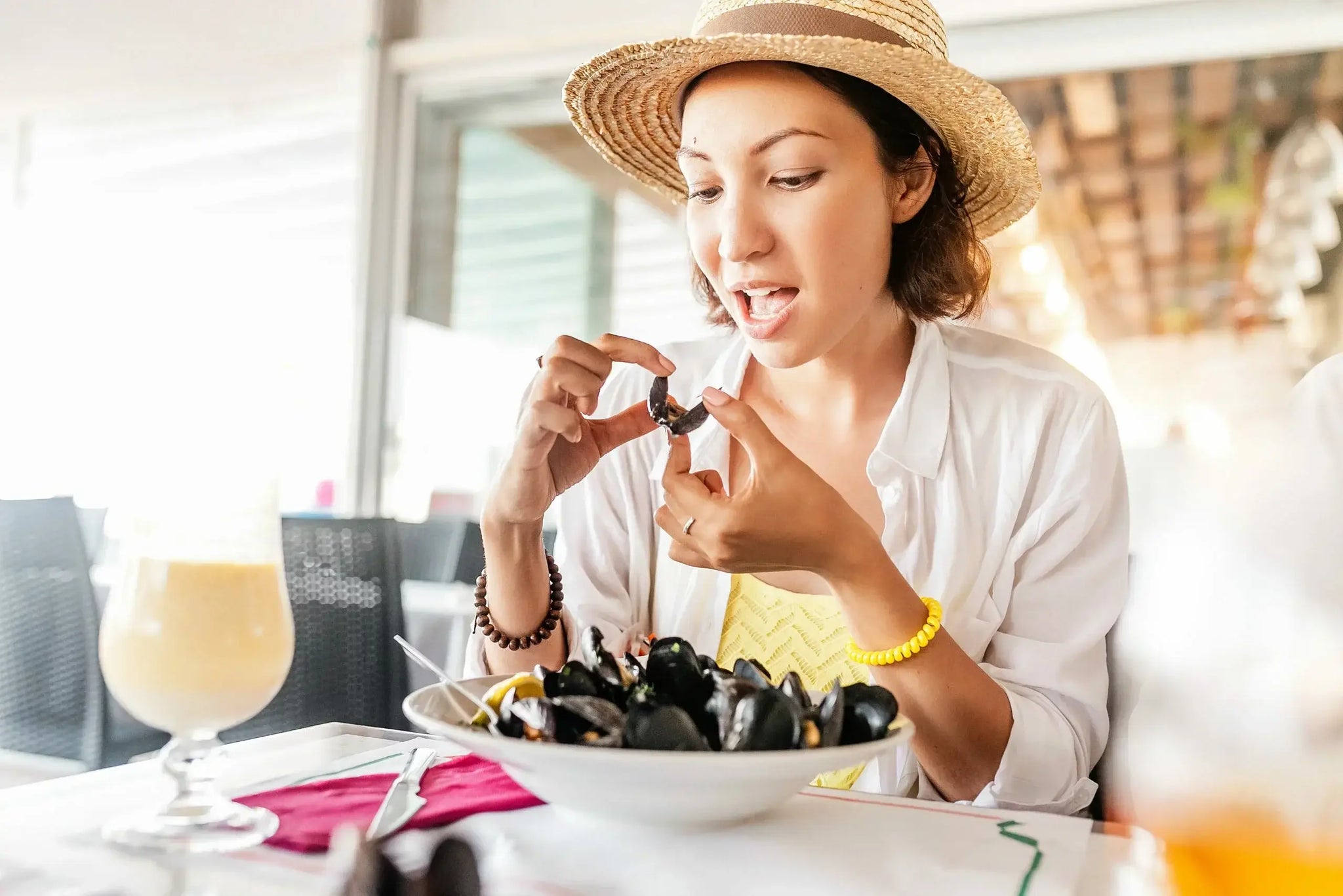 A woman wearing a straw hat and white shirt is seated at a table in a restaurant, eating mussels. Are mussels vegan