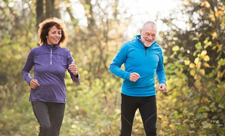 A man and a woman are jogging outdoors on a sunny day, embracing one of the 4 Ways to Keep Running After 50. The woman wears a purple jacket and the man a blue one, both smiling and soaking in the greenery surrounding them.