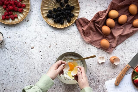 A person in a green shirt is cracking an egg into a bowl with flour on a speckled countertop.
