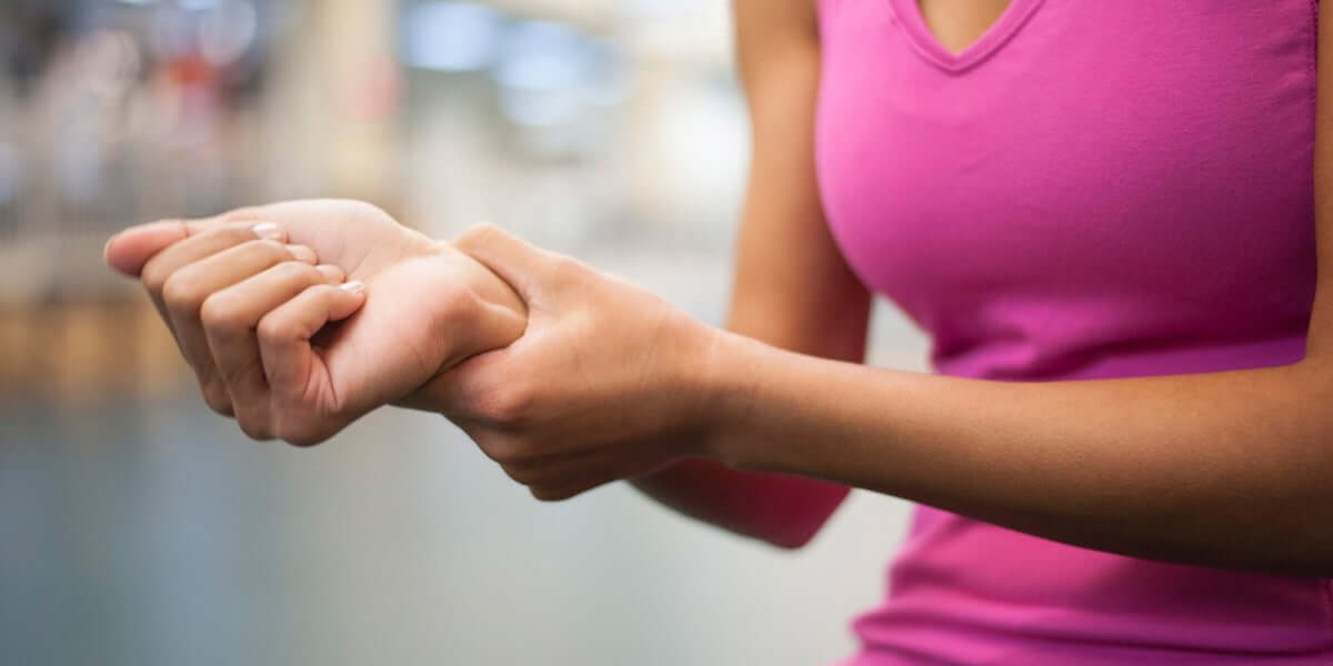 A person wearing a pink tank top is holding their wrist with the opposite hand, suggesting an injury or pain in the wrist.