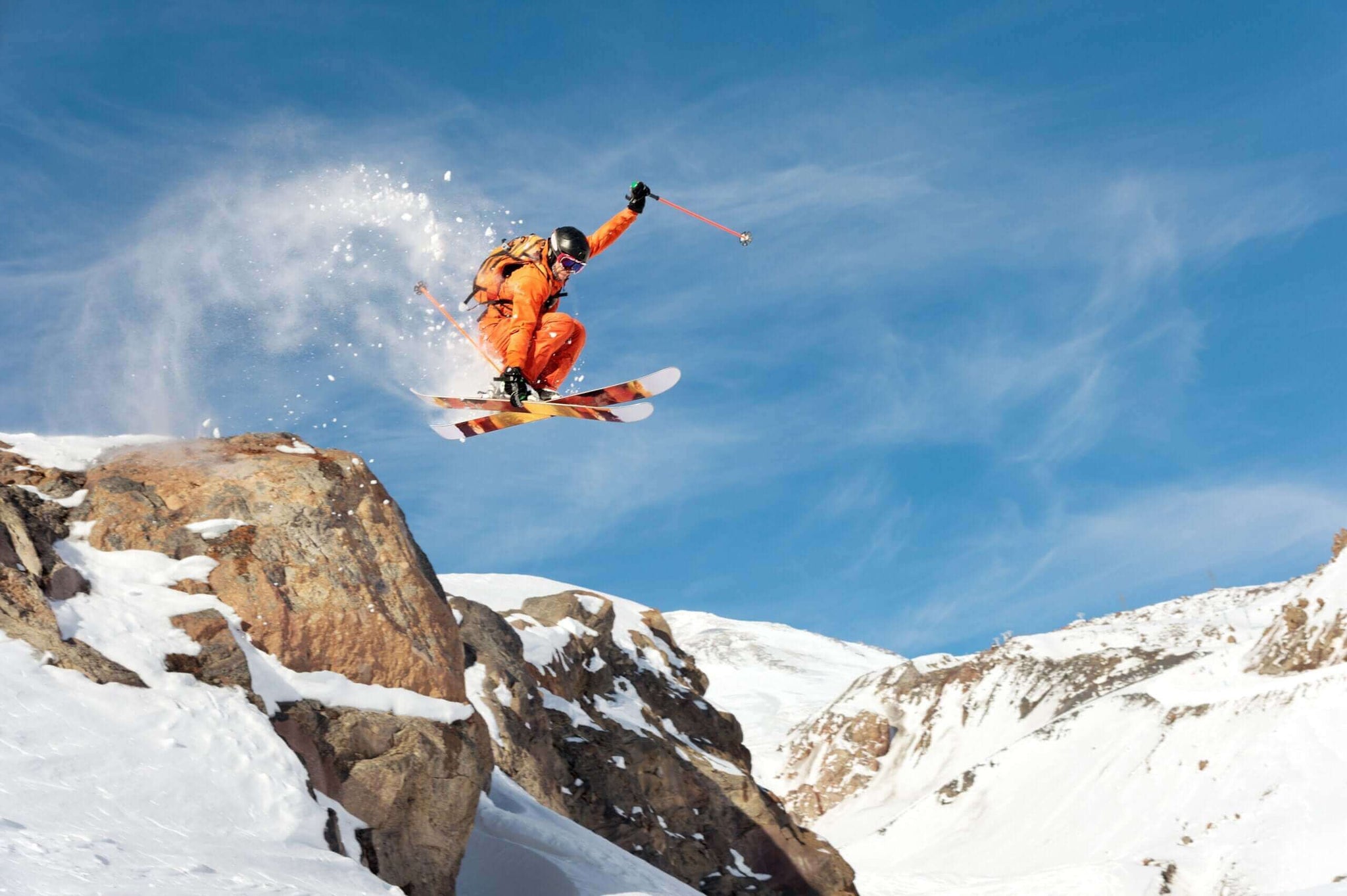 A skier in an orange outfit performs a jump off a rocky ledge on a snowy mountain, with snow spraying around them. Action sports