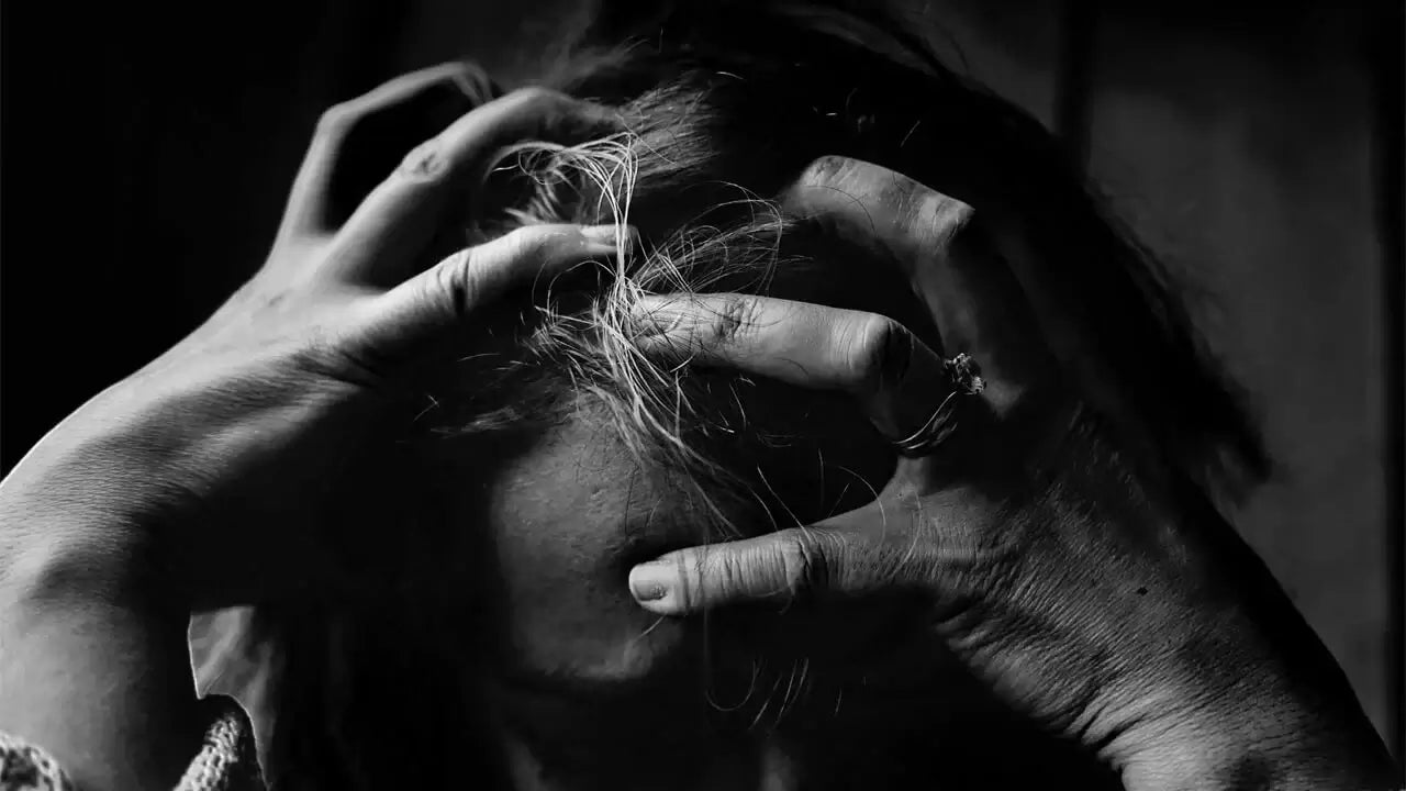 A black and white photo of a person with long hair, grasping their head with both hands in a distressed manner.