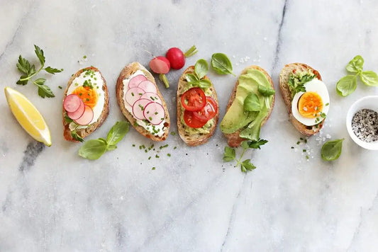 A row of five toasted bread slices topped with various ingredients: a hard-boiled egg and radish, radish slices, tomato and greens, avocado slices