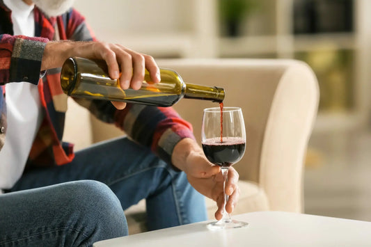 A person is sitting on a couch, wearing a red plaid shirt and blue jeans, and pouring red wine from a bottle into a wine glass.