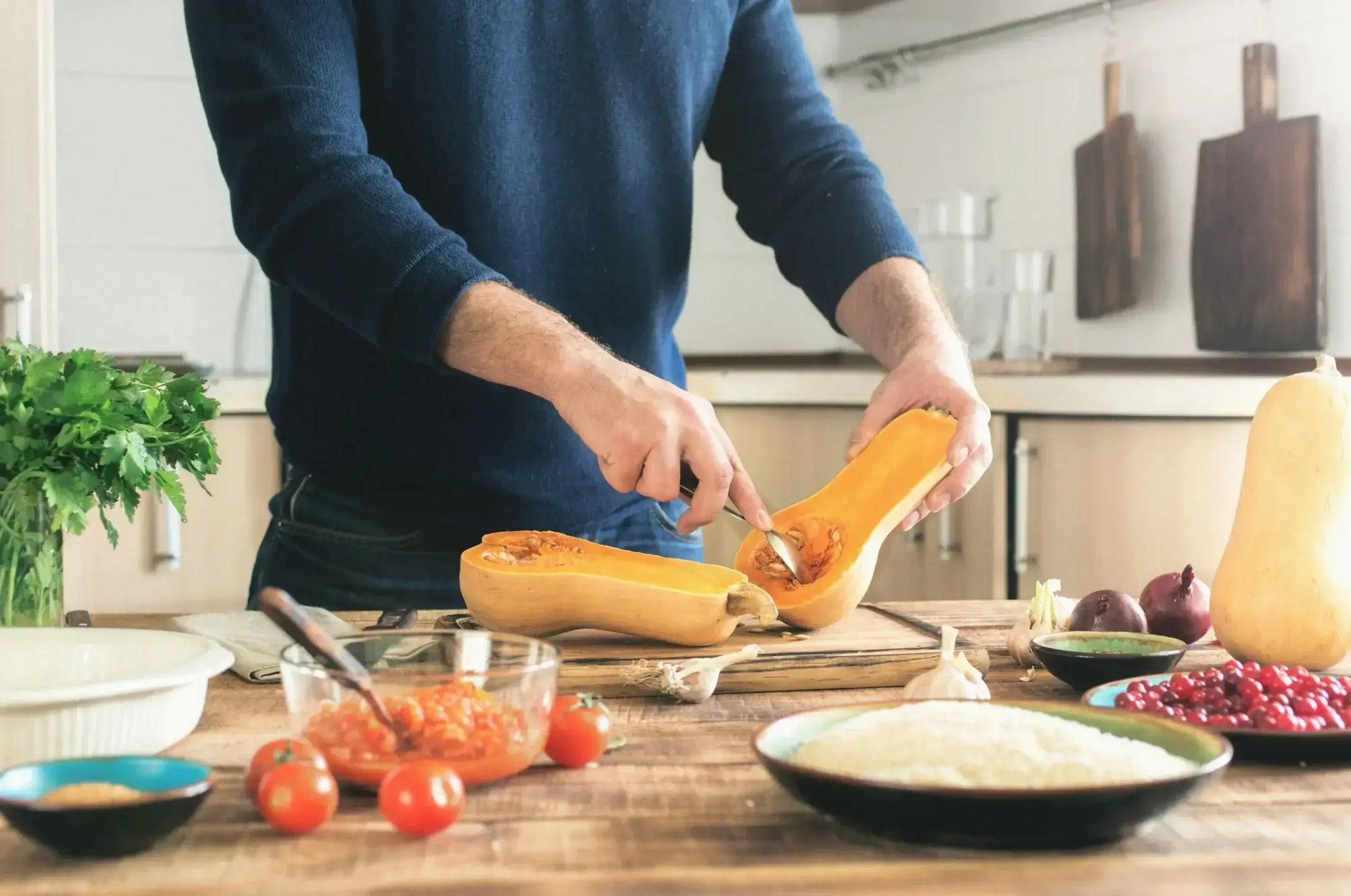 A person in a dark blue sweater is cutting a butternut squash in half on a wooden countertop. Use that fall