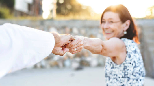 A smiling woman with dark hair and a patterned dress holds hands with another person,
