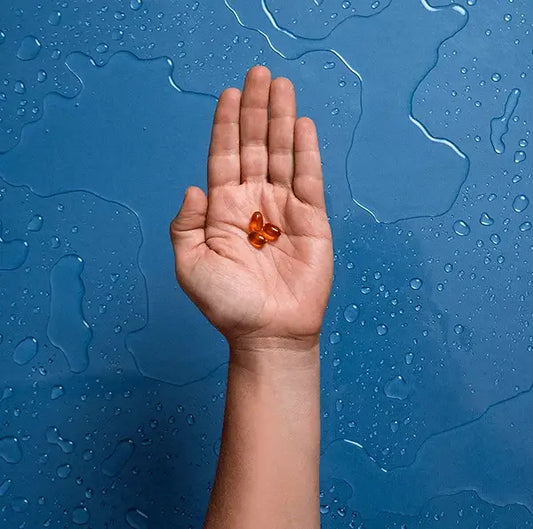 A hand is held up against a blue background with water droplets. The open palm contains three small, brown, oval-shaped capsules or pills, prompting one to wonder how much green lipped mussel should you take for optimal benefits.