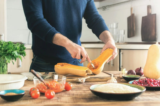 A person in a dark blue sweater is cutting a butternut squash in half on a wooden countertop.
