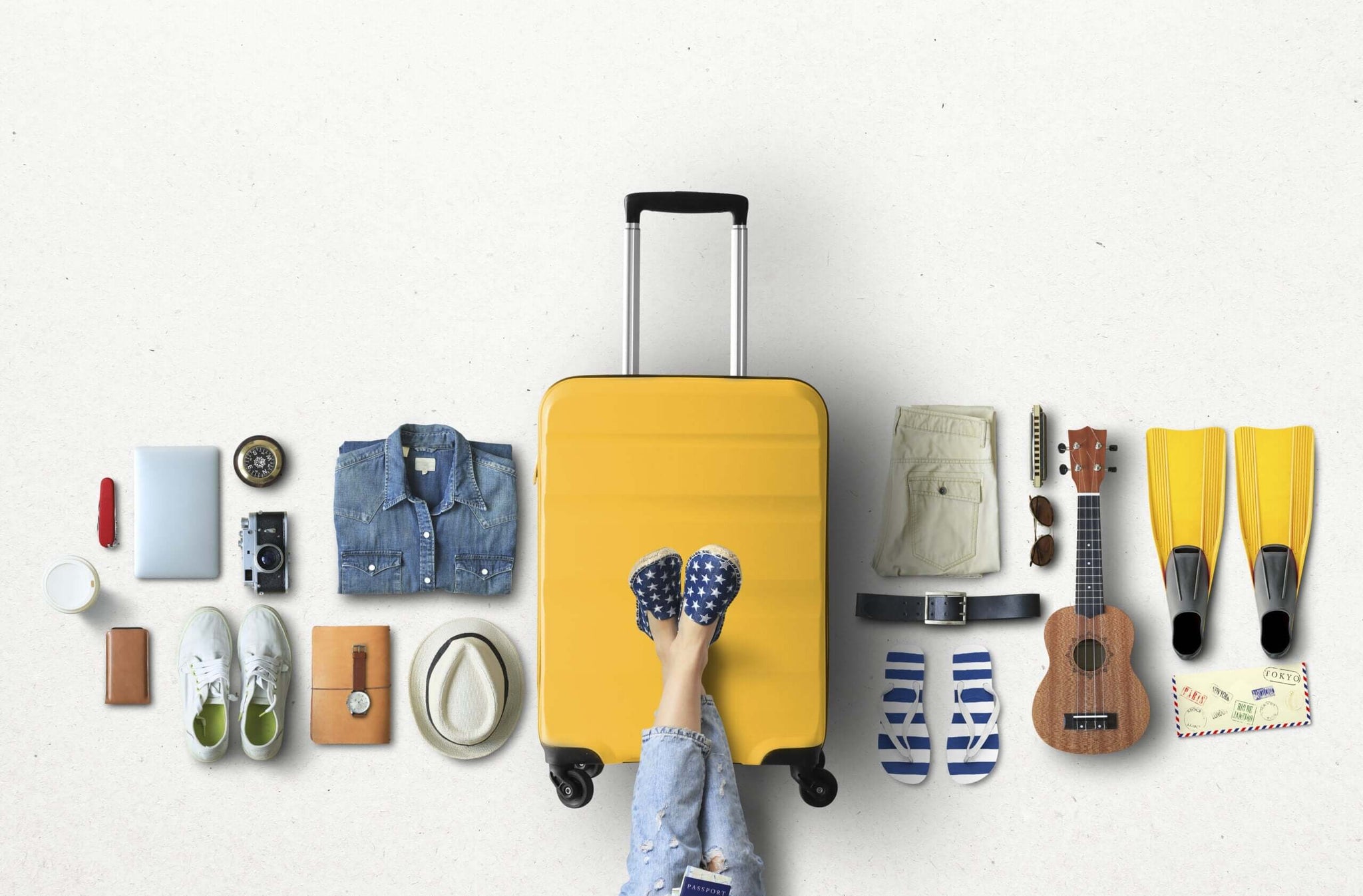 Overhead view of various travel items neatly arranged around a central yellow suitcase.