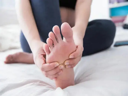 A person sitting on a bed is shown massaging the sole of their bare foot with both hands, indicating they might be dealing with gout signs symptoms and treatments you can’t ignore.