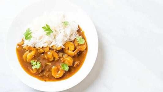 A white plate containing shrimp curry with a rich brown sauce and garnished with cilantro is served alongside a generous portion of steamed white rice.