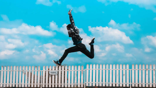 A person in dark clothing is captured mid-air, jumping over a white picket fence against a backdrop of a bright blue sky with scattered clouds.