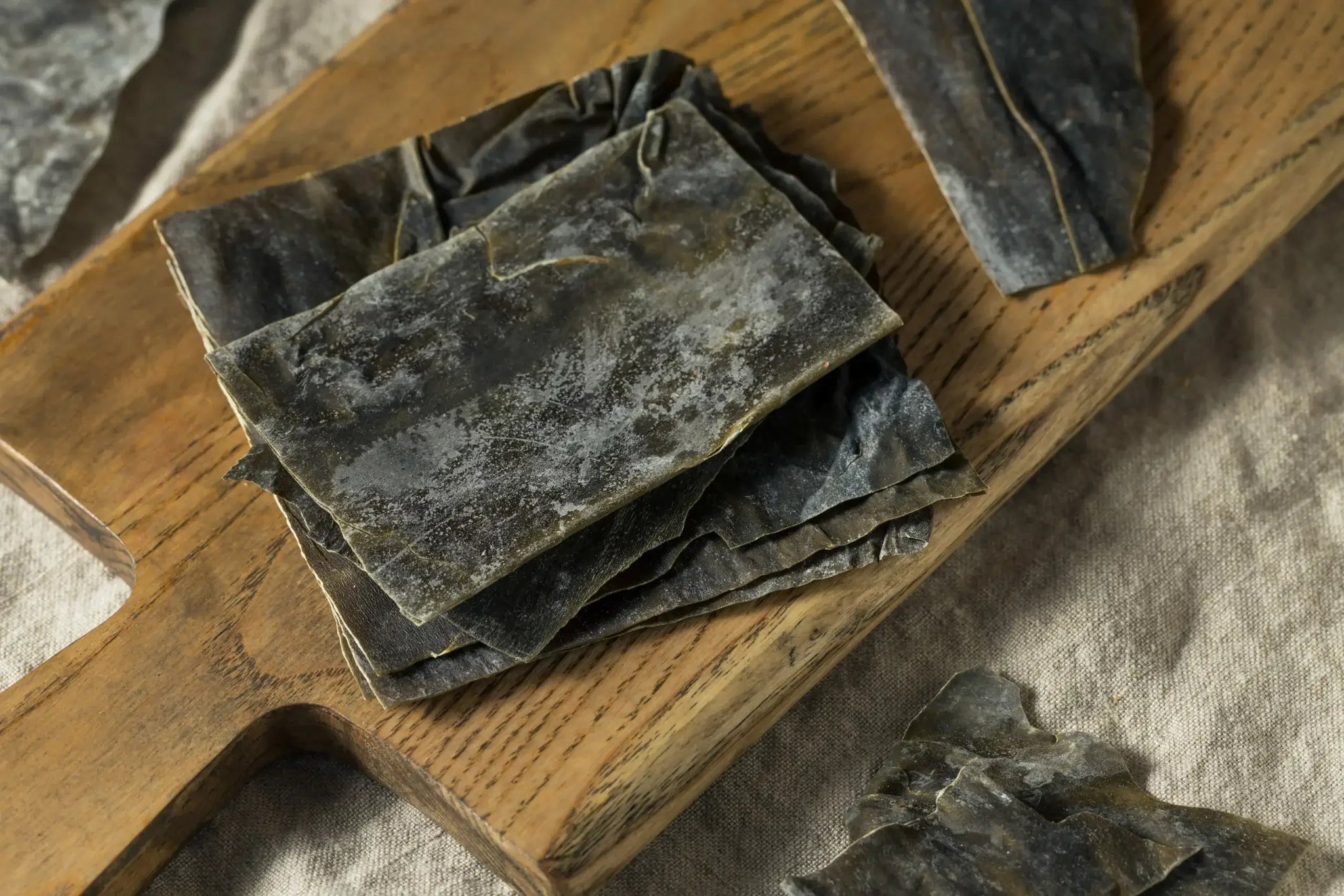 Dried kelp sheets stacked on a wooden cutting board with some pieces scattered around on a light fabric surface.