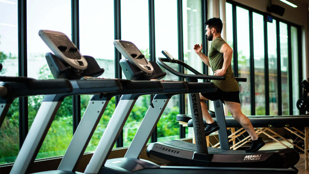 A man with a beard is running on a treadmill in a gym with large windows, embracing natural ways to strengthen bones and joints.