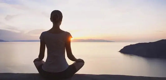 A person sits in a cross-legged position, facing a serene body of water during sunset.