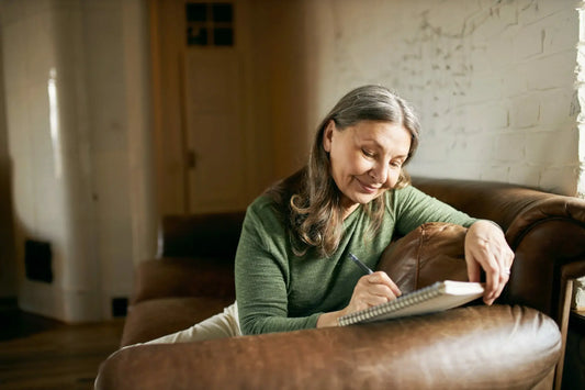 A woman with long gray hair sits comfortably on a brown leather sofa, writing in a notebook with a content expression.
