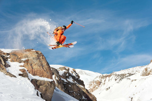 A skier in an orange outfit performs a jump off a rocky ledge on a snowy mountain, with snow spraying around them.