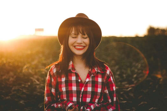 A woman with long dark hair and red lipstick stands in a field at sunset, smiling with her eyes closed.