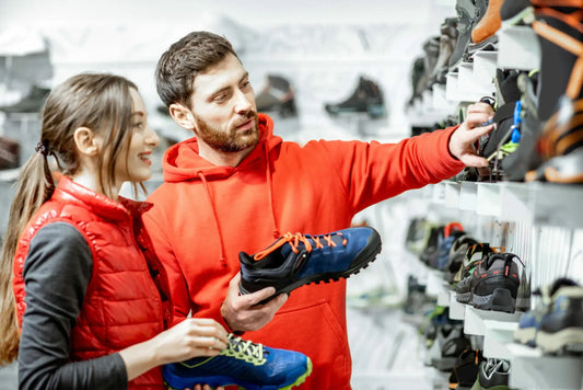 A man in a red hoodie and a woman in a red vest are shopping for hiking shoes.