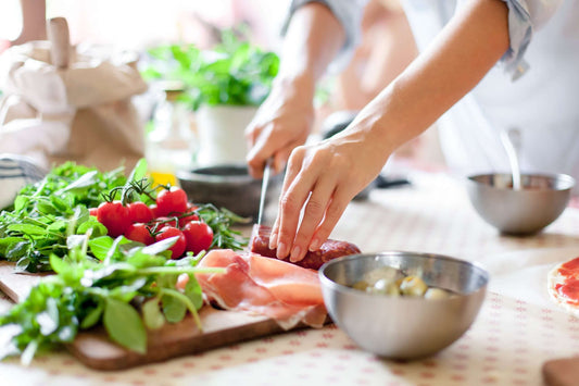 A person slicing meat on a cutting board surrounded by various fresh ingredients, including cherry tomatoes, herbs, and leafy greens.