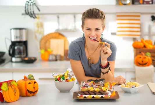 A woman with pulled-back hair is smiling and eating a snack in a kitchen decorated for Halloween.