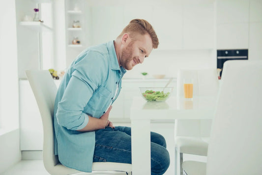 A man with red hair and a beard, wearing a light blue shirt and jeans, sits at a white kitchen table