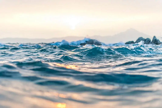 Close-up of gentle ocean waves with sunlight reflecting off the surface. The sky is pale and hazy with the sun low on the horizon.