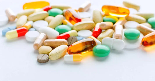 A variety of colorful pills and capsules are scattered on a white surface. The assortment includes round tablets, oval capsules