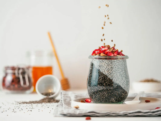 A glass jar filled with chia pudding is topped with pomegranate seeds and nuts, with some of the toppings being sprinkled over it.