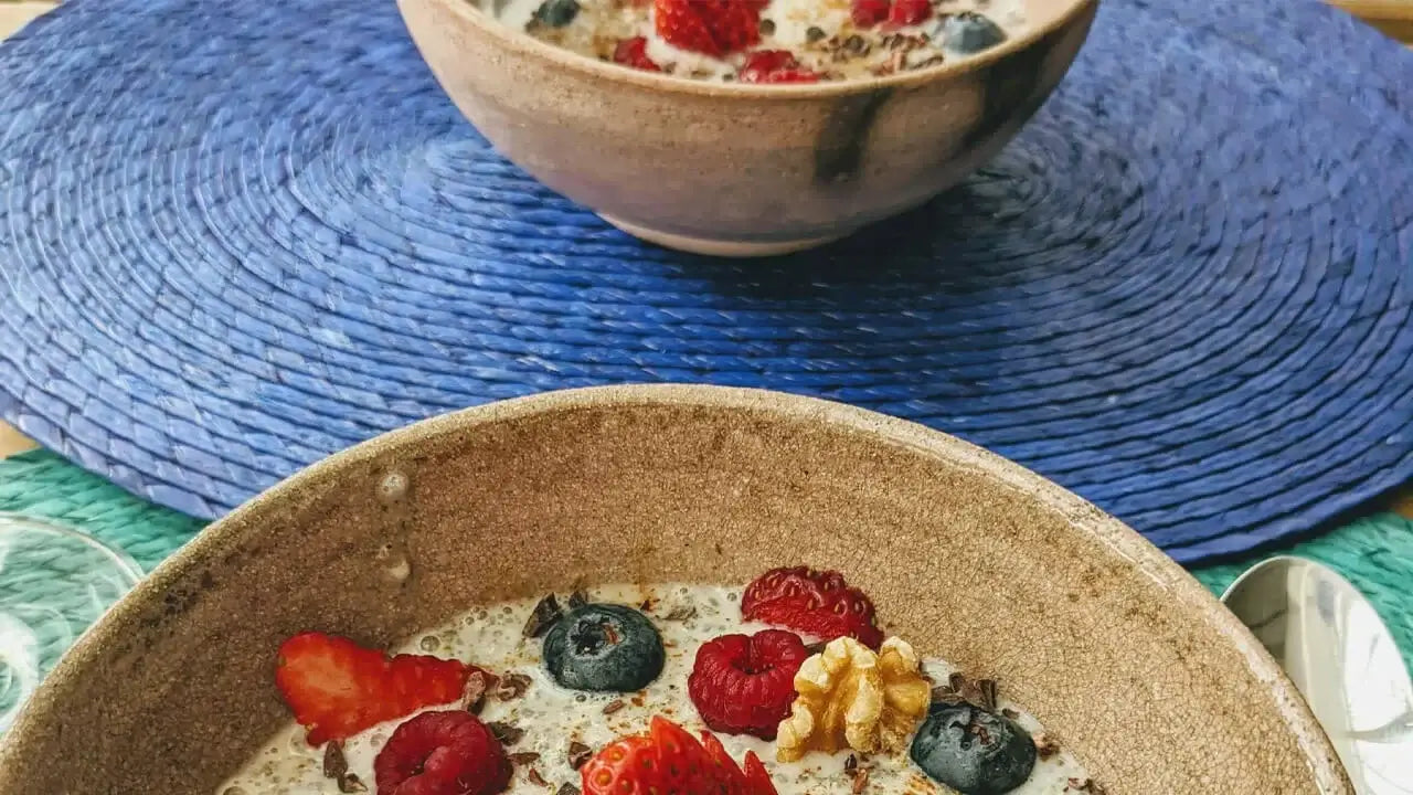 Two bowls of oatmeal topped with fresh strawberries, blueberries, raspberries, and a walnut piece are placed on a blue woven mat. \