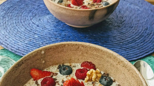 Two bowls of oatmeal topped with fresh strawberries, blueberries, raspberries, and a walnut piece are placed on a blue woven mat. \