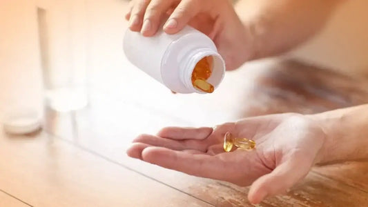 A person is holding and pouring yellow capsule pills from a white bottle into their hand. The scene is set on a wooden table with a glass of water in the background, suggesting they might be considering Omega-3