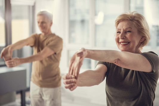 Two senior individuals, one man and one woman, are smiling and stretching their arms forward in a bright room.