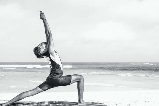 A person wearing athletic clothing performs a yoga pose, stretching one arm forward and the other upward on a beach. The background features the sea and a cloudy sky. The pose emphasizes balance, strength, and flexibility.
