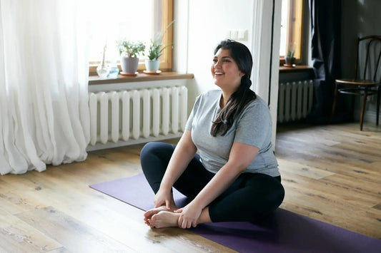 A woman sits on a purple yoga mat in a butterfly stretch position, smiling and looking relaxed. Post-exercise