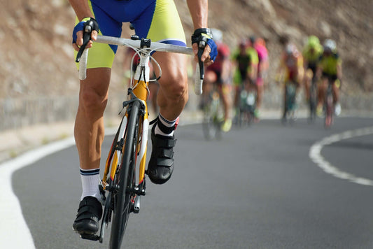 Close-up of a cyclist wearing yellow and blue spandex, black shoes, and gloves, gripping the handlebars while riding on a paved road.