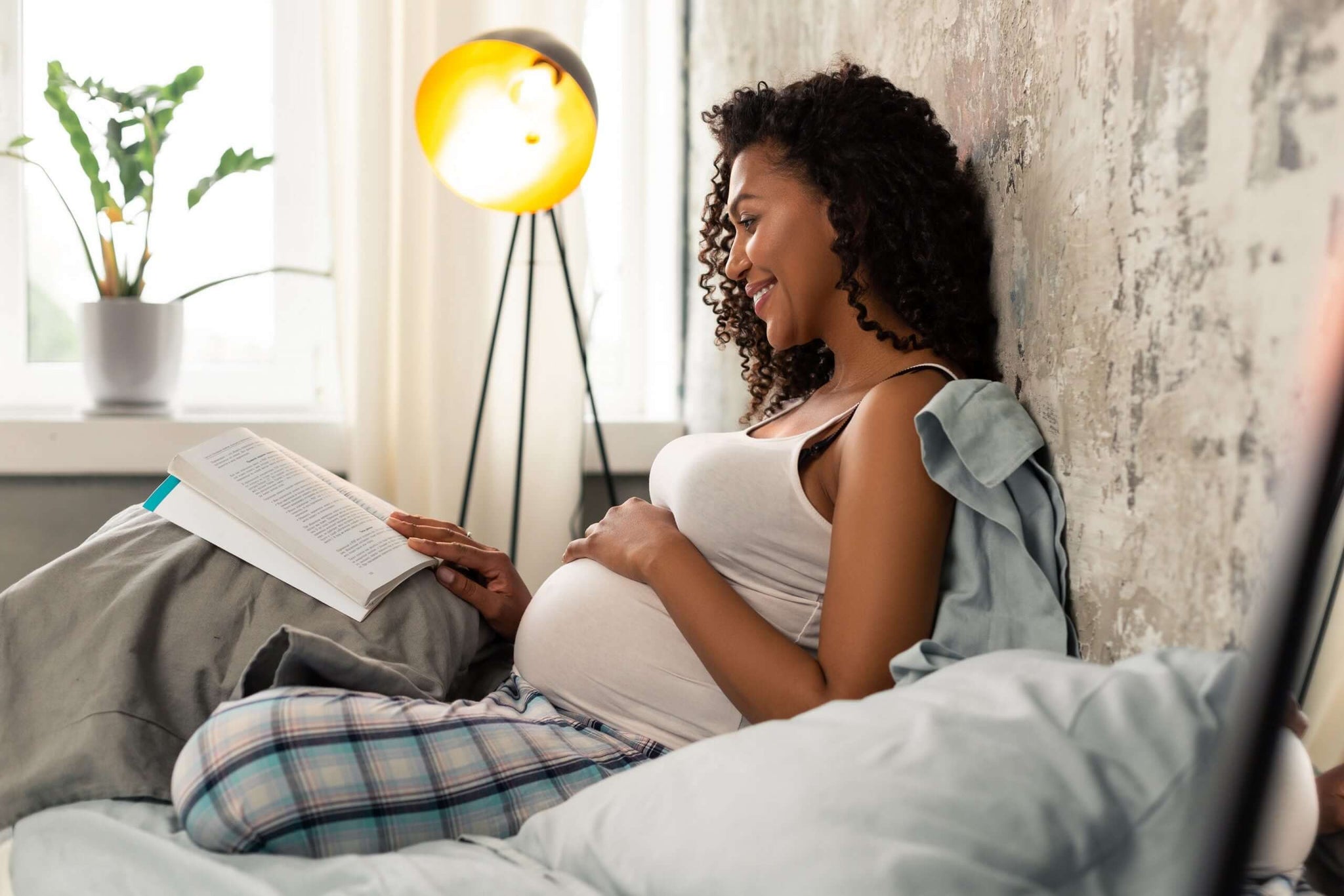 A smiling pregnant woman sits on a cozy bed, reading a book.