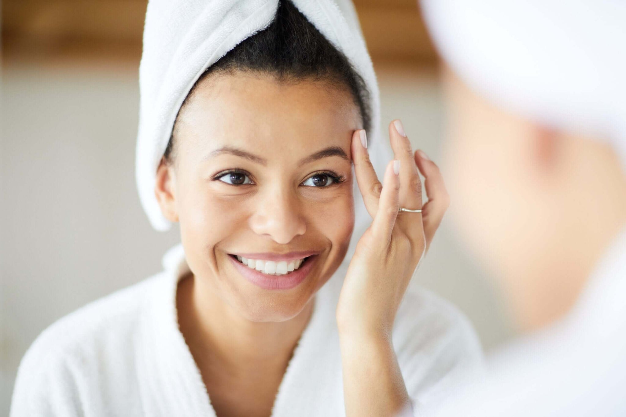 A smiling woman with a towel wrapped around her head stands in front of a mirror, touching her face gently.