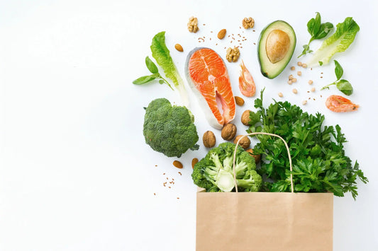 A brown paper bag on a white background, overflowing with healthy foods, including broccoli, salmon, avocado, shrimp